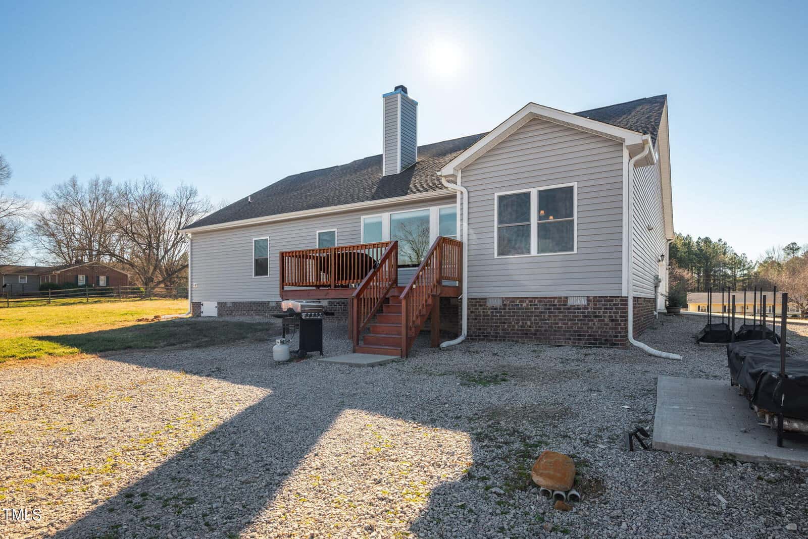 A modern two-story house with beige vinyl and brick siding features a wooden deck with stairs leading to the backyard. there's a bright sun overhead and gravel around the house's perimeter.