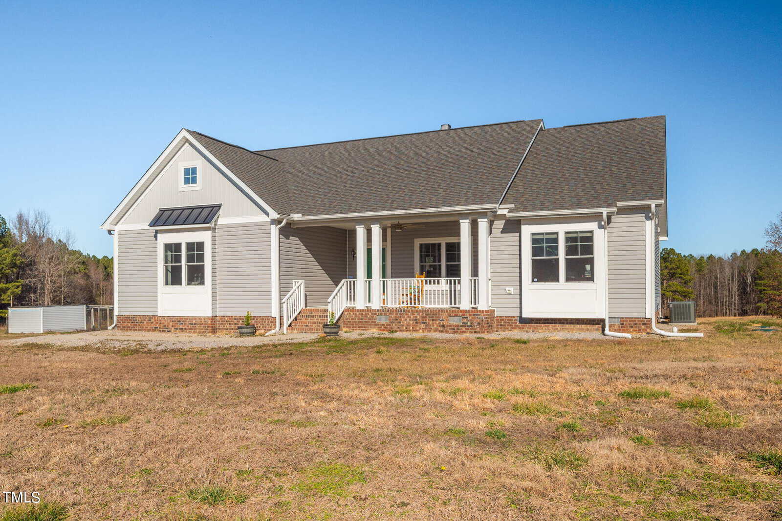 A single-story suburban home with a large front porch, tan siding, and a dark shingled roof. the house is situated in a grassy field with a clear blue sky above.