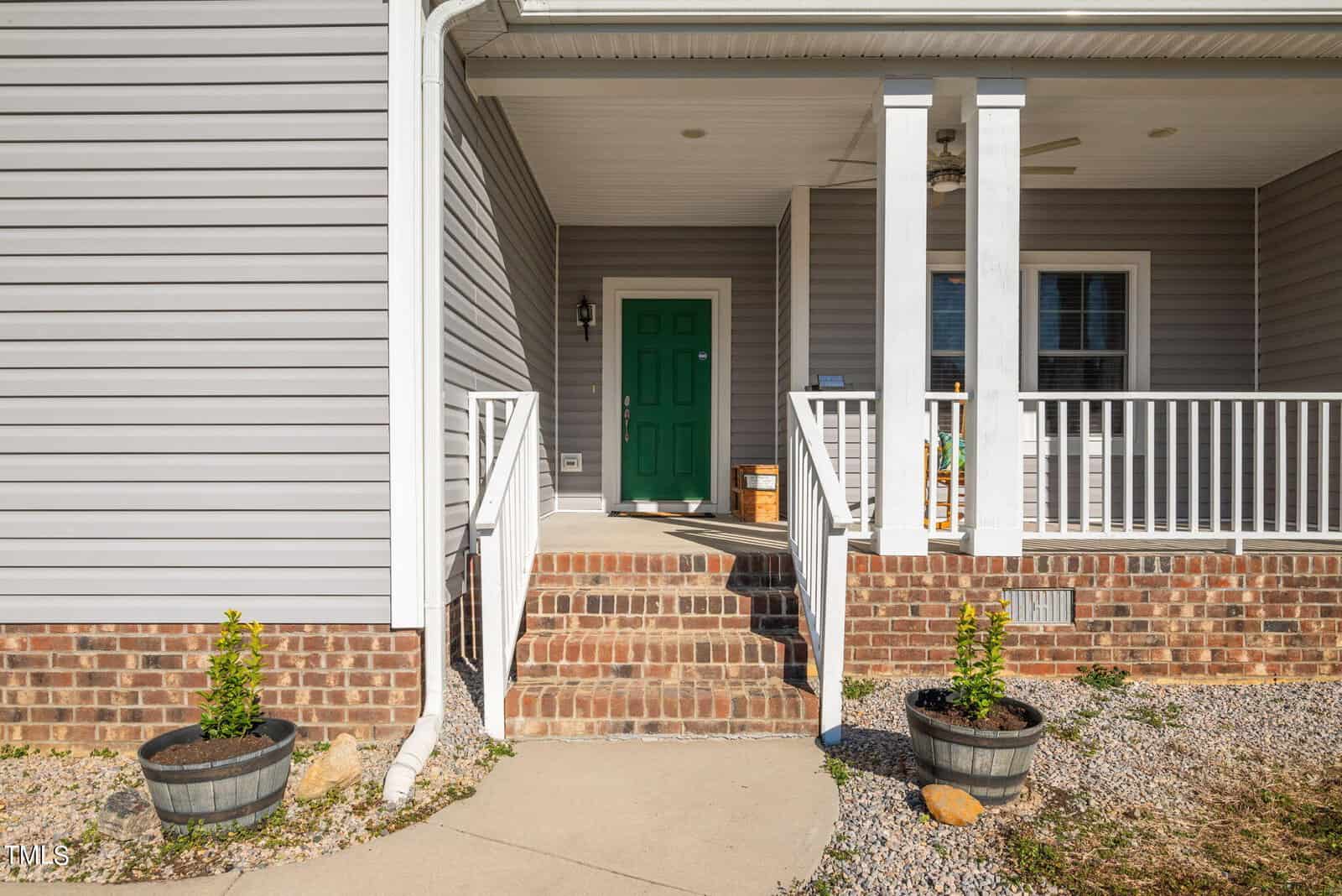Front view of a house with brick stairs leading to a porch with white railings, featuring a bright green door, under a clear sky. two potted plants are by the steps.