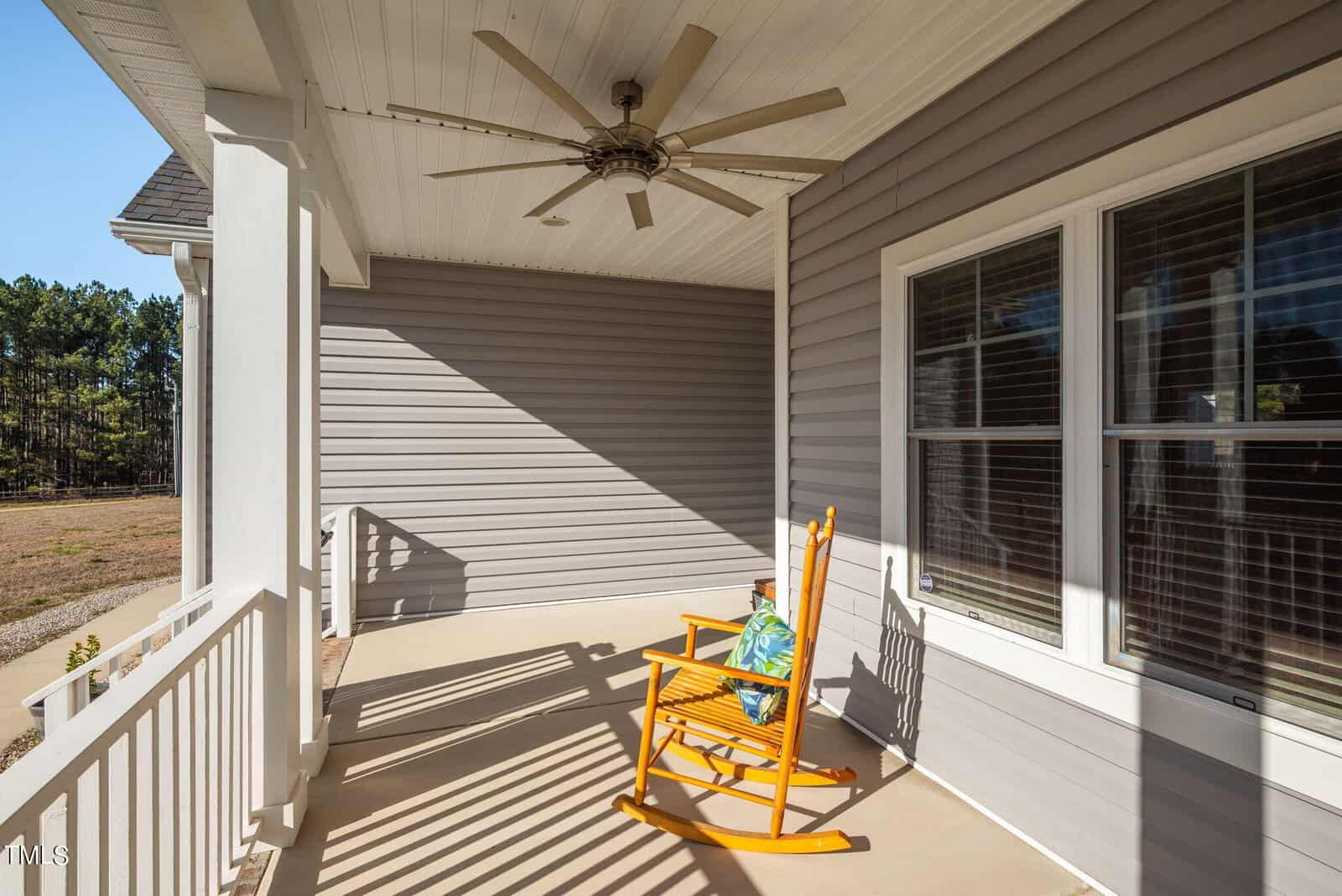 A sunny porch with a rocking chair, featuring a ceiling fan overhead and views of a grassy yard through windows and the side railing.