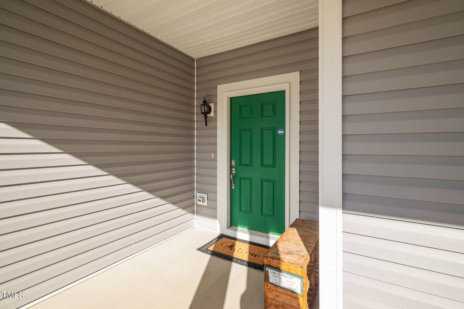 A vibrant green door on a house with gray siding, illuminated by sunlight with a shadow pattern across the porch. a small lantern-style light fixture hangs beside the door.