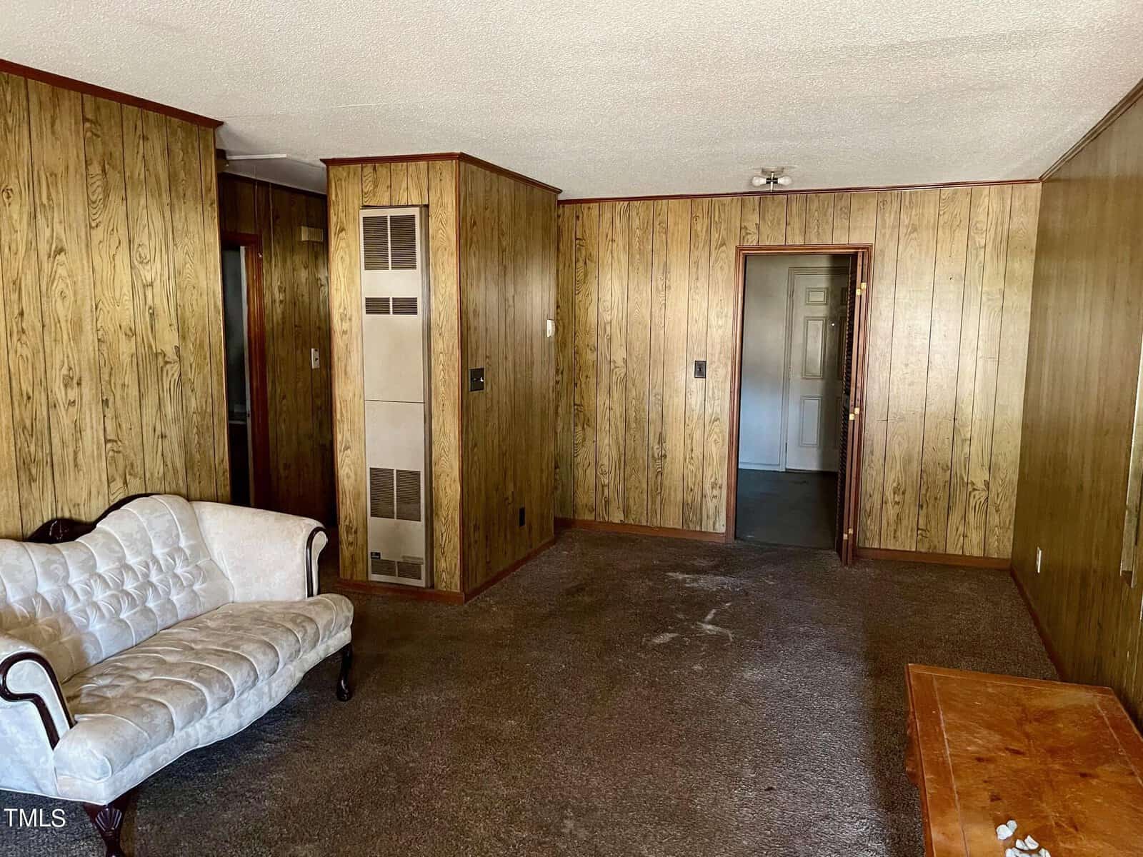 A living room with wood paneling, a white sofa, a wooden coffee table, and brown carpeting, showing signs of wear.