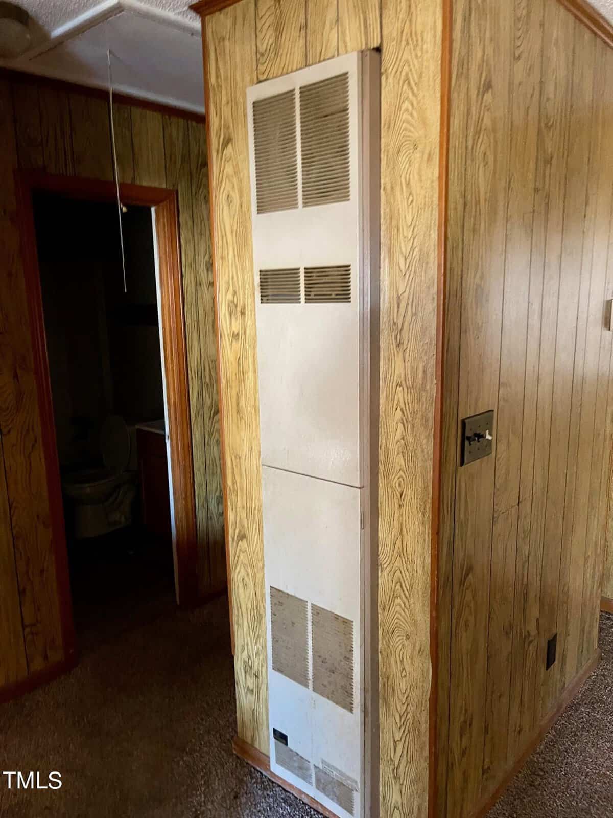 Interior of a room with wood paneling, showing a dated hvac wall unit and a glimpse of a bathroom through an open door.