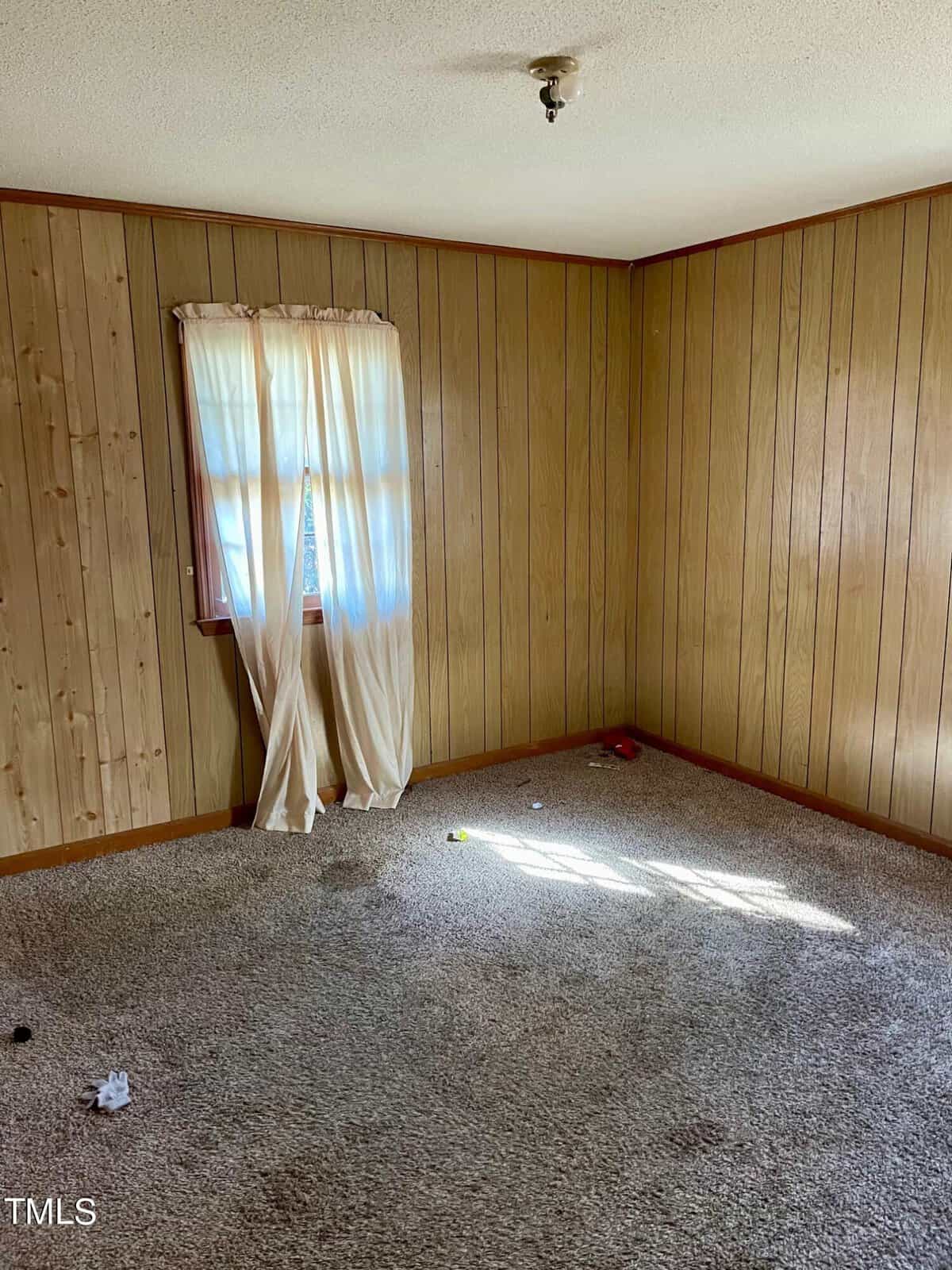 An empty room with wood paneling, a single window with sheer curtains, and a beige carpet with a few small items scattered on the floor. sunlight streams in through the window, casting a bright patch on the carpet.