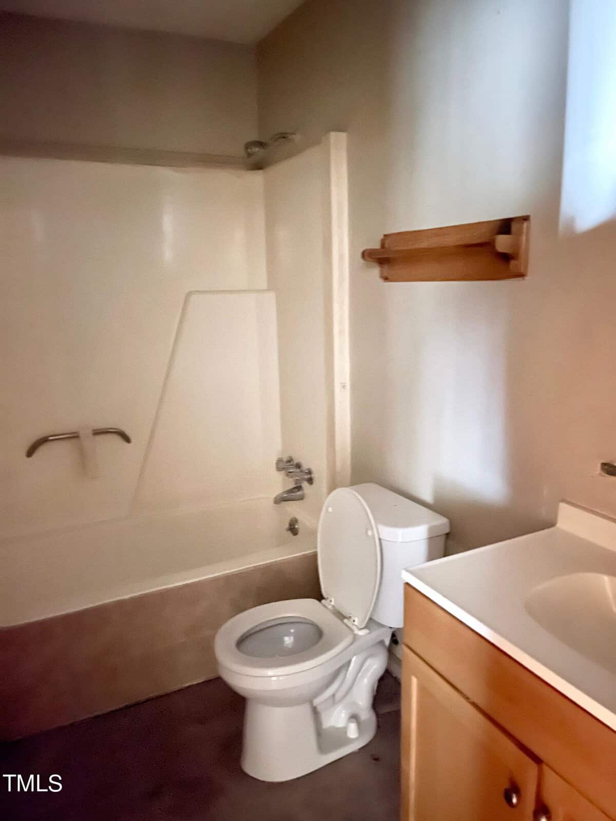 A modest bathroom featuring a toilet, bathtub with shower fixtures, and a wooden shelf, all under warm lighting.