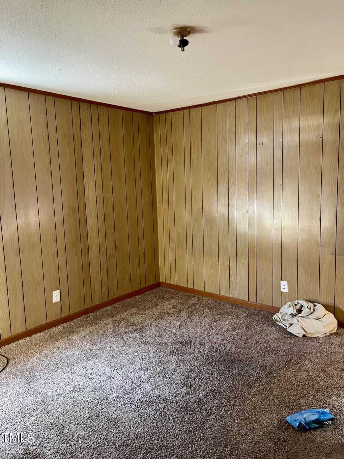 An empty room with wood paneling on the walls, a shaggy brown carpet on the floor, and a small pile of clothes in the corner.