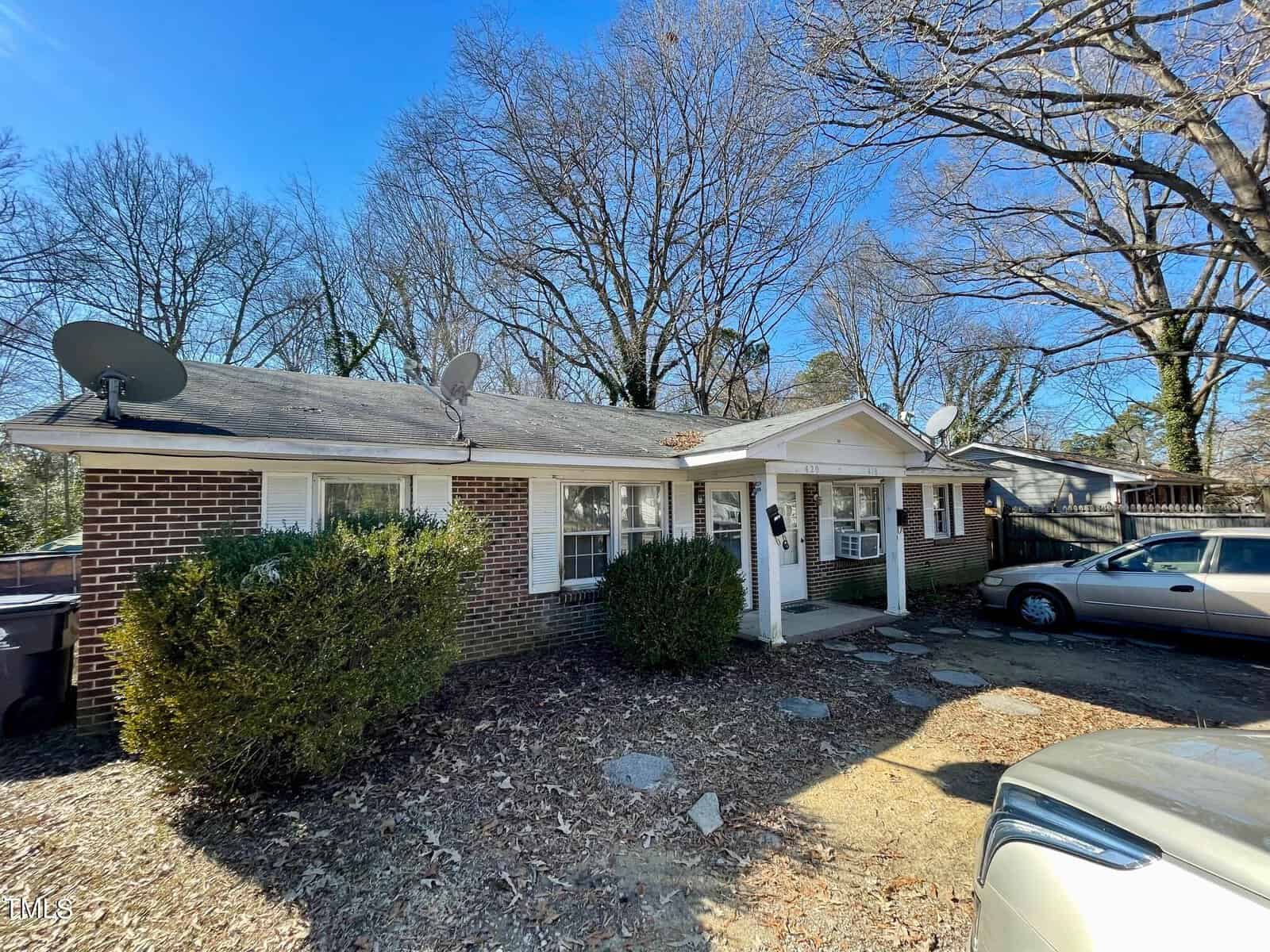 A modest single-story brick house with a white door and trim, situated at 218/220 Mitchell Street in Henderson, NC, features two satellite dishes on the roof. It is surrounded by bare trees