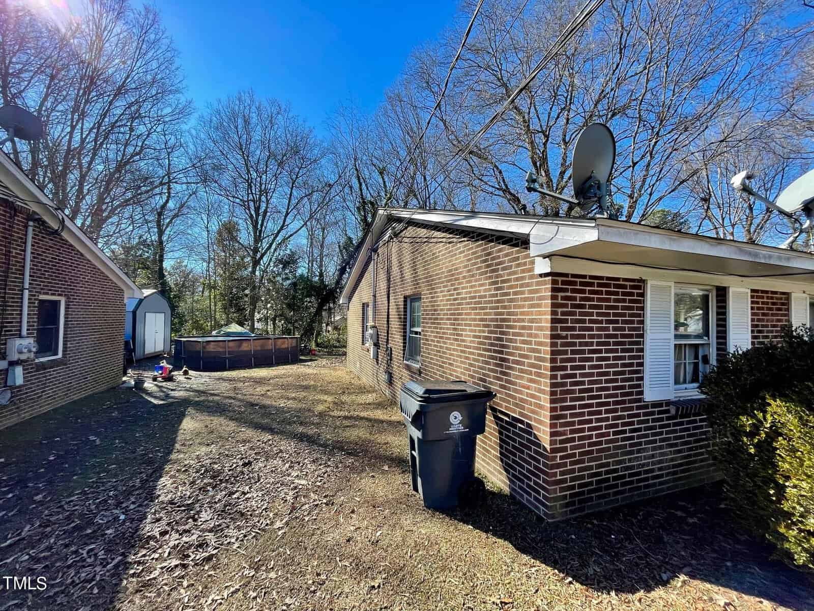 A brick suburban house on a sunny day with a clear sky, featuring satellite dishes and trash bins on the side, and a fenced backyard with trees in the background.