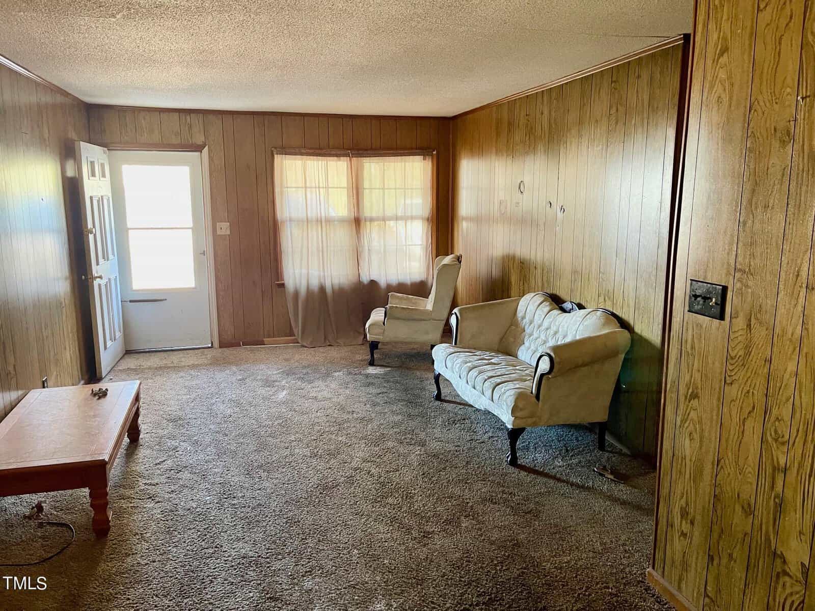 An interior view of a room with wood paneling on the walls, furnished with a cream-colored couch and chair, a coffee table, and carpeted flooring. a window with curtains and a closed door are visible on the far side of the room.