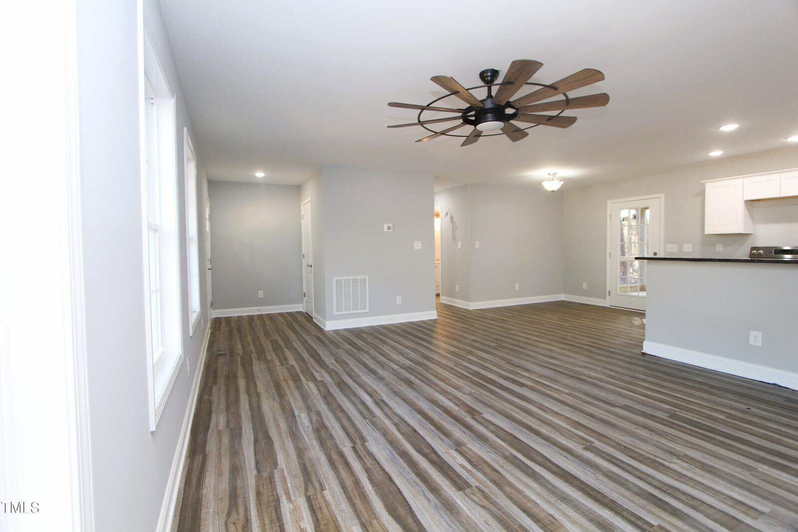 Spacious room with light gray walls and wood flooring. Features a large ceiling fan with wooden blades, recessed lighting, and a partially visible kitchen area with white cabinets and a breakfast bar. Multiple windows allow natural light.