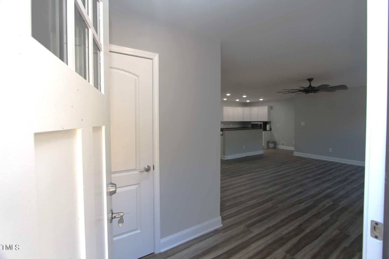 View of a bright, modern interior from the entrance. The room features light gray walls, wood flooring, and a ceiling fan. The open layout includes a kitchen with bar-style seating and recessed lighting in the ceiling.