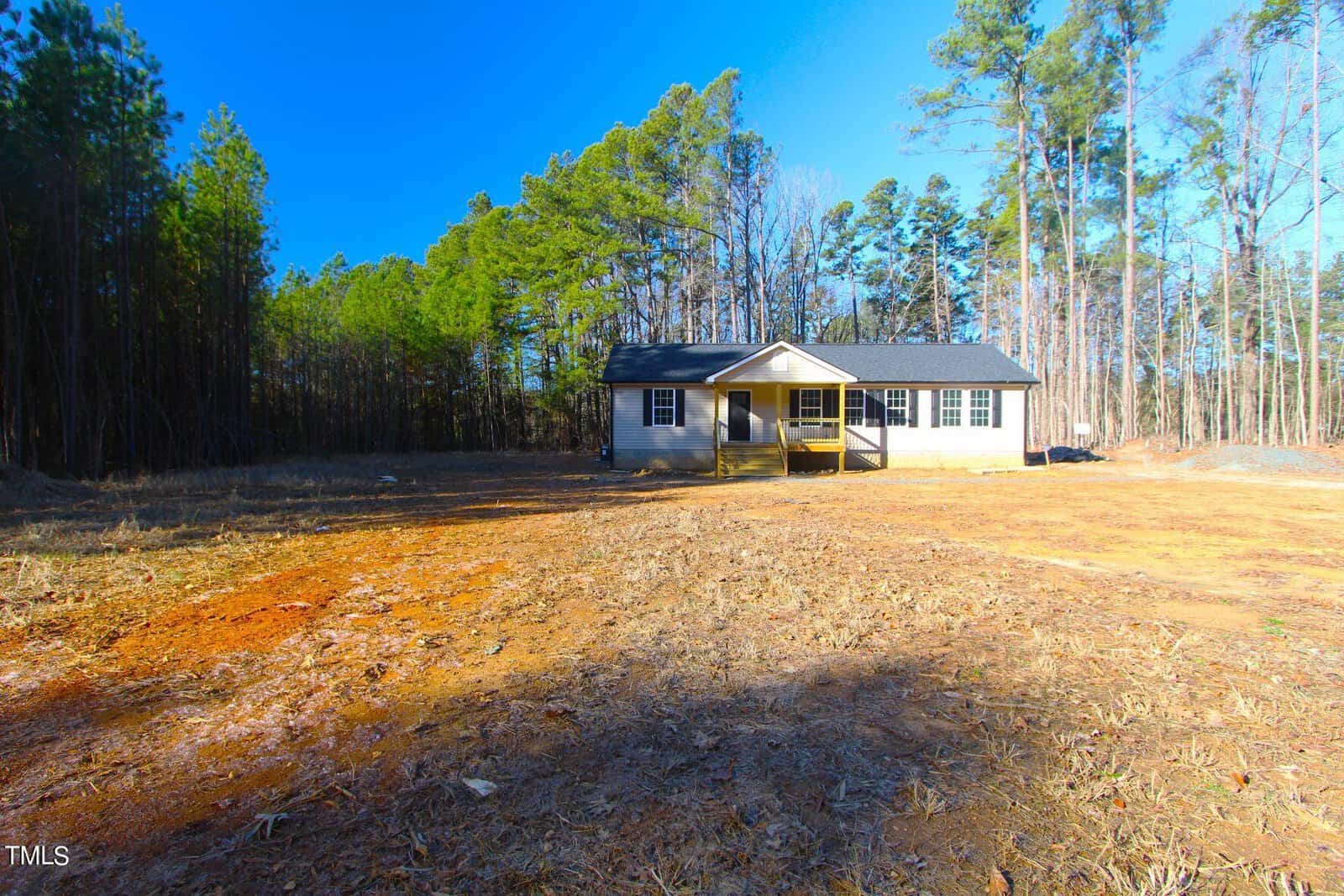 A small, single-story house with a porch is situated in a clearing surrounded by tall trees. The ground is bare and reddish, and the sky is clear and blue.