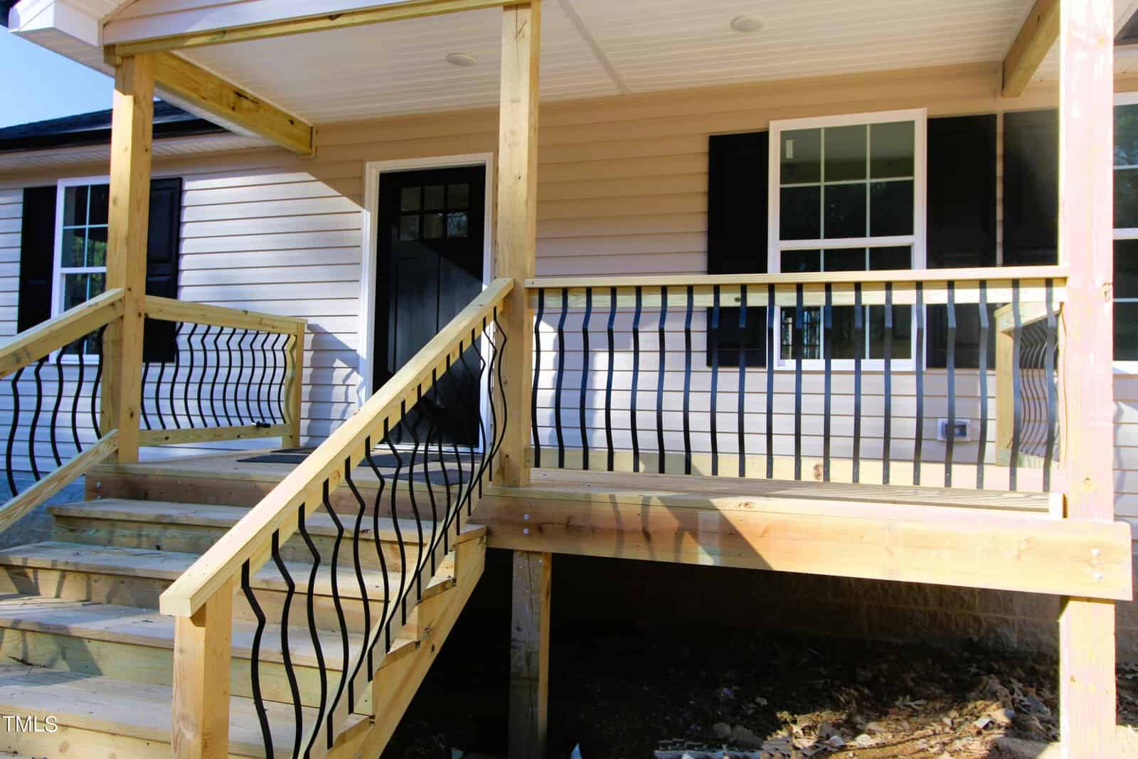Wooden porch and stairway with black metal railings attached to a beige house. The porch is partially shaded, leading to a black front door. Two windows with black shutters are visible on the house exterior.