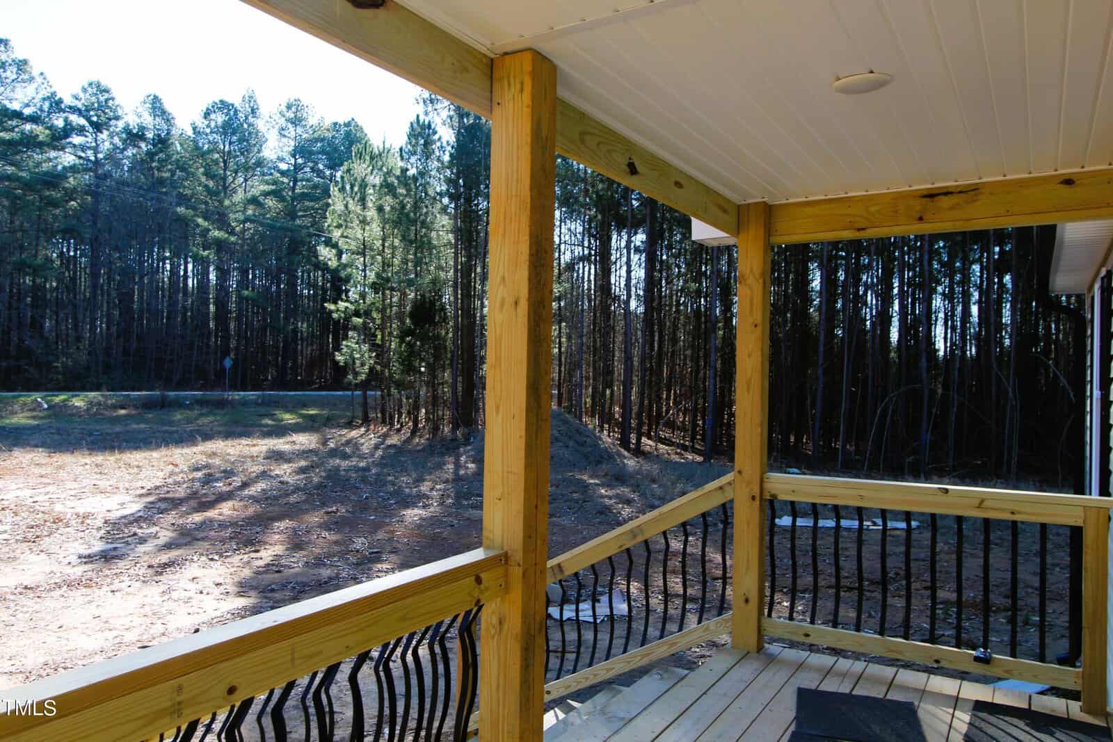 View from a wooden porch overlooking a clearing surrounded by dense trees. The porch has sturdy wooden posts and a railing with black spindles. The sun casts shadows on the ground, highlighting the natural setting.