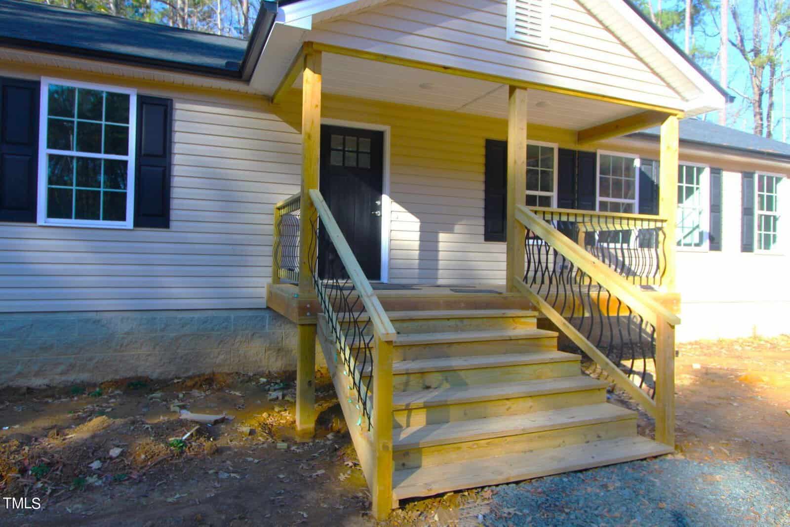 A small front porch of a beige house with wooden stairs and railing. The porch has a black front door and is supported by wooden pillars. Sunlight casts shadows on the siding, surrounded by bare ground and debris.
