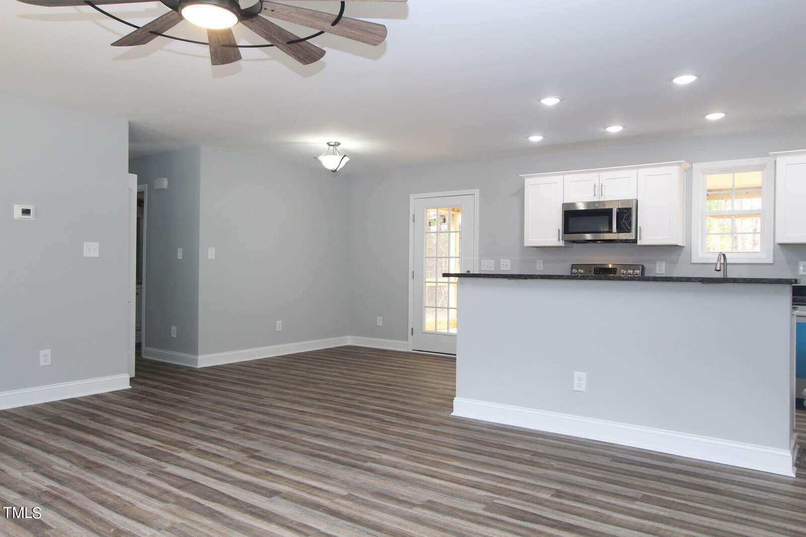 A modern kitchen and dining area with light gray walls and wood-style flooring. The kitchen features white cabinets, a stainless steel microwave, and recessed lighting. A ceiling fan is visible, and a door with glass panels leads outside.