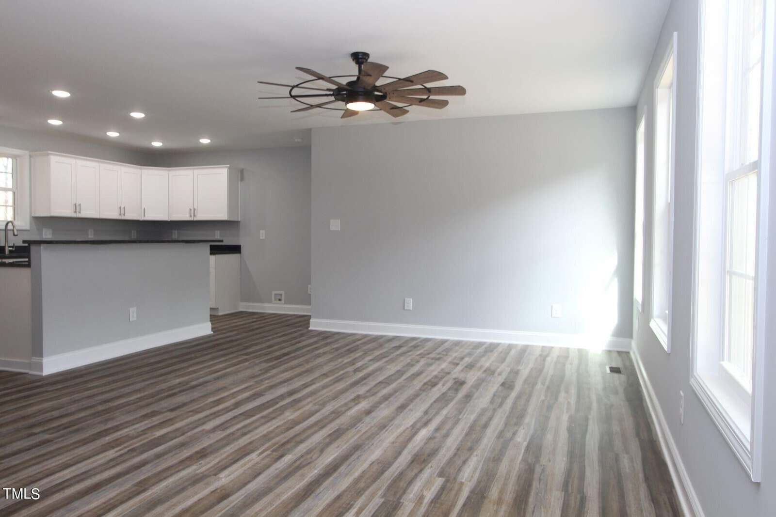 A modern, empty living room with gray walls and wood-patterned flooring. A ceiling fan with wooden blades hangs above. White cabinets and a black countertop are visible in the adjoining kitchen area on the left. Large windows allow natural light in.