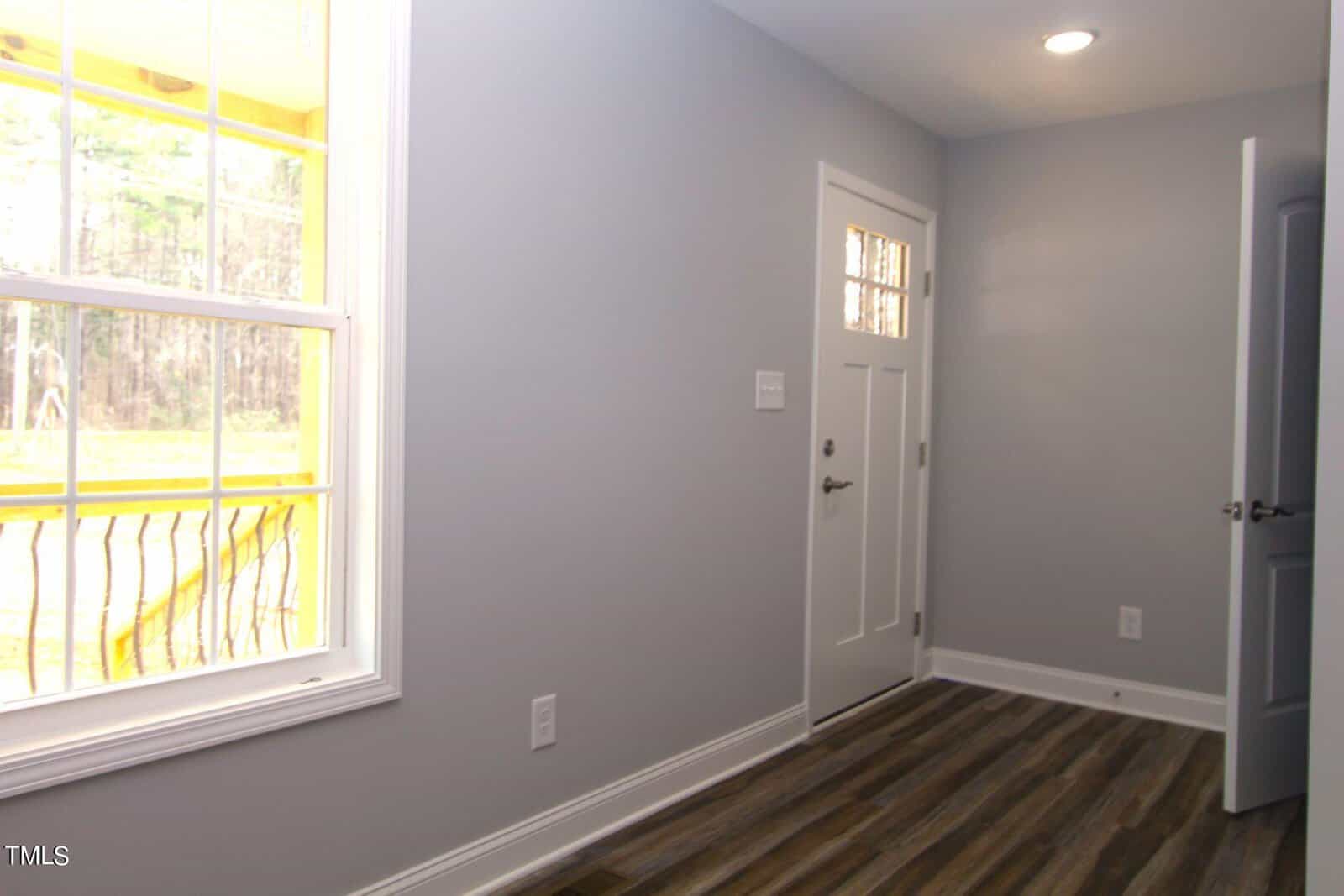 A small foyer with a white entrance door and wood-style flooring. A large window on the left lets in natural light, and the walls are painted light gray. A wooden railing is visible outside through the window.