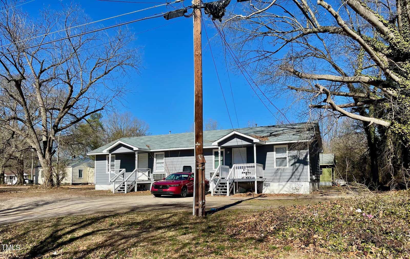 A single-story house with a gray exterior and white trim featuring a front porch with stairs, parked next to a red car, on a sunny day with trees and a utility pole in the foreground.