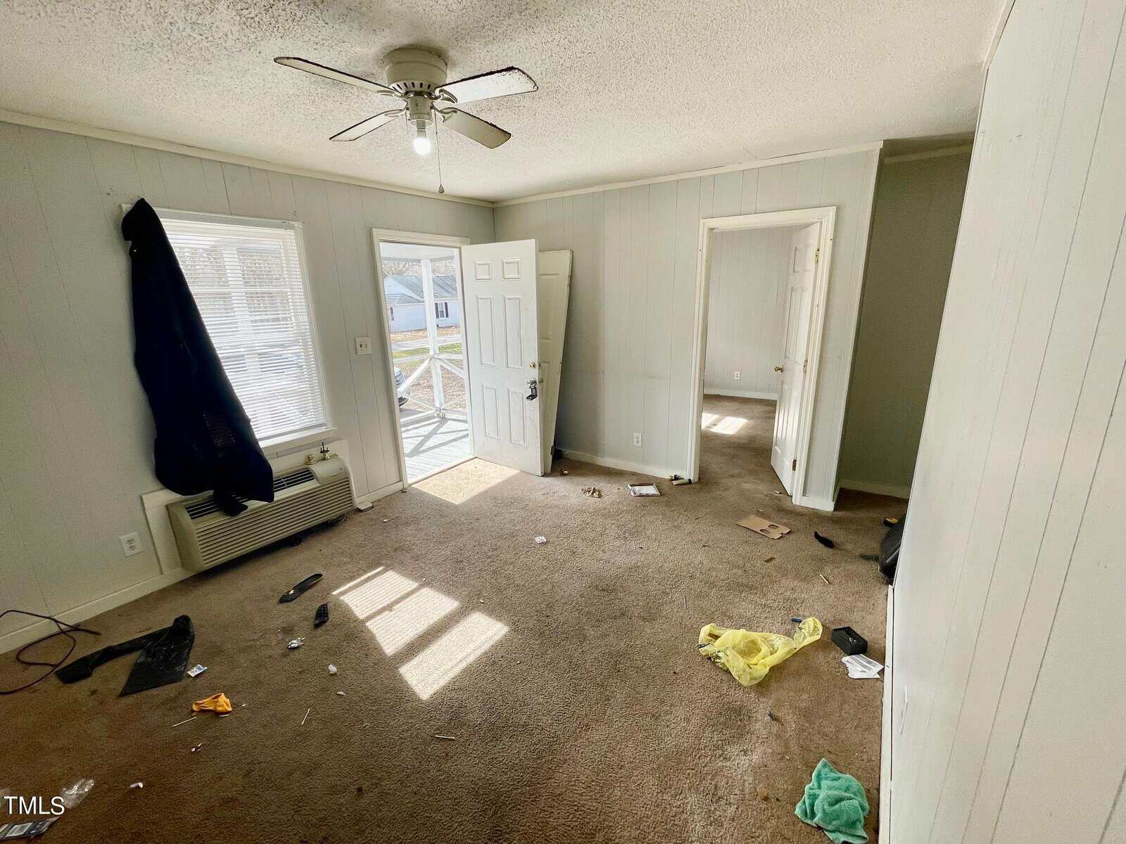 An empty room with scattered debris on the carpet, a ceiling fan overhead, and sunlight streaming through a window next to an open door.