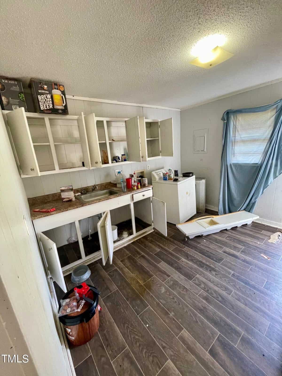 A cluttered, unfinished kitchen with open cabinets, assorted items on the counter, a disheveled curtain covering a window, and a wooden plank floor.