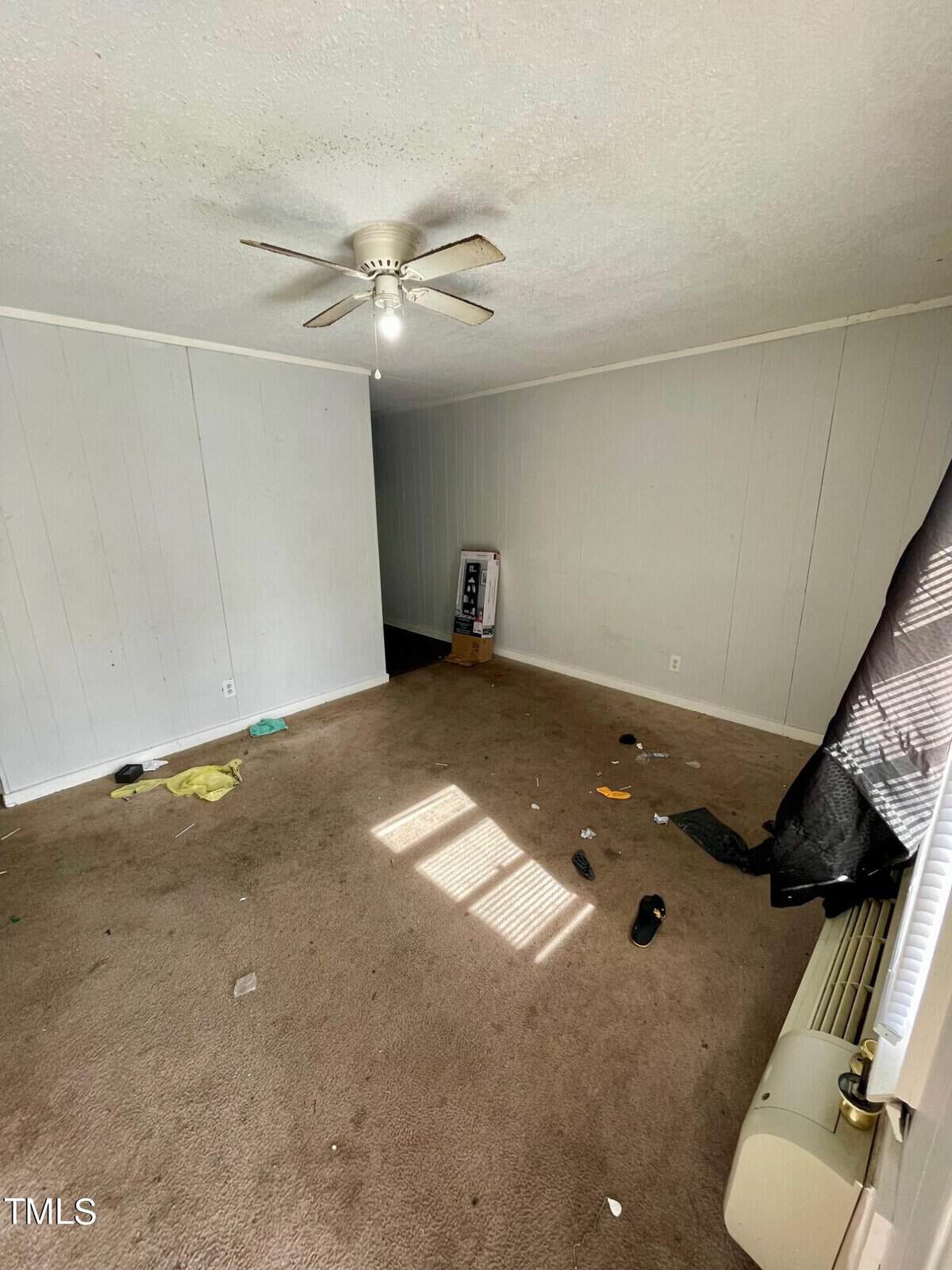 An empty room with beige carpet, white walls, and a ceiling fan, showing some discarded items on the floor and natural light coming in through the window.