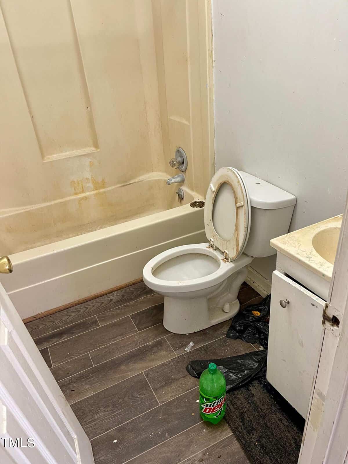 A neglected bathroom showing a dirty toilet, a stained bathtub, and debris on the floor, with a plastic bottle left beside the toilet.