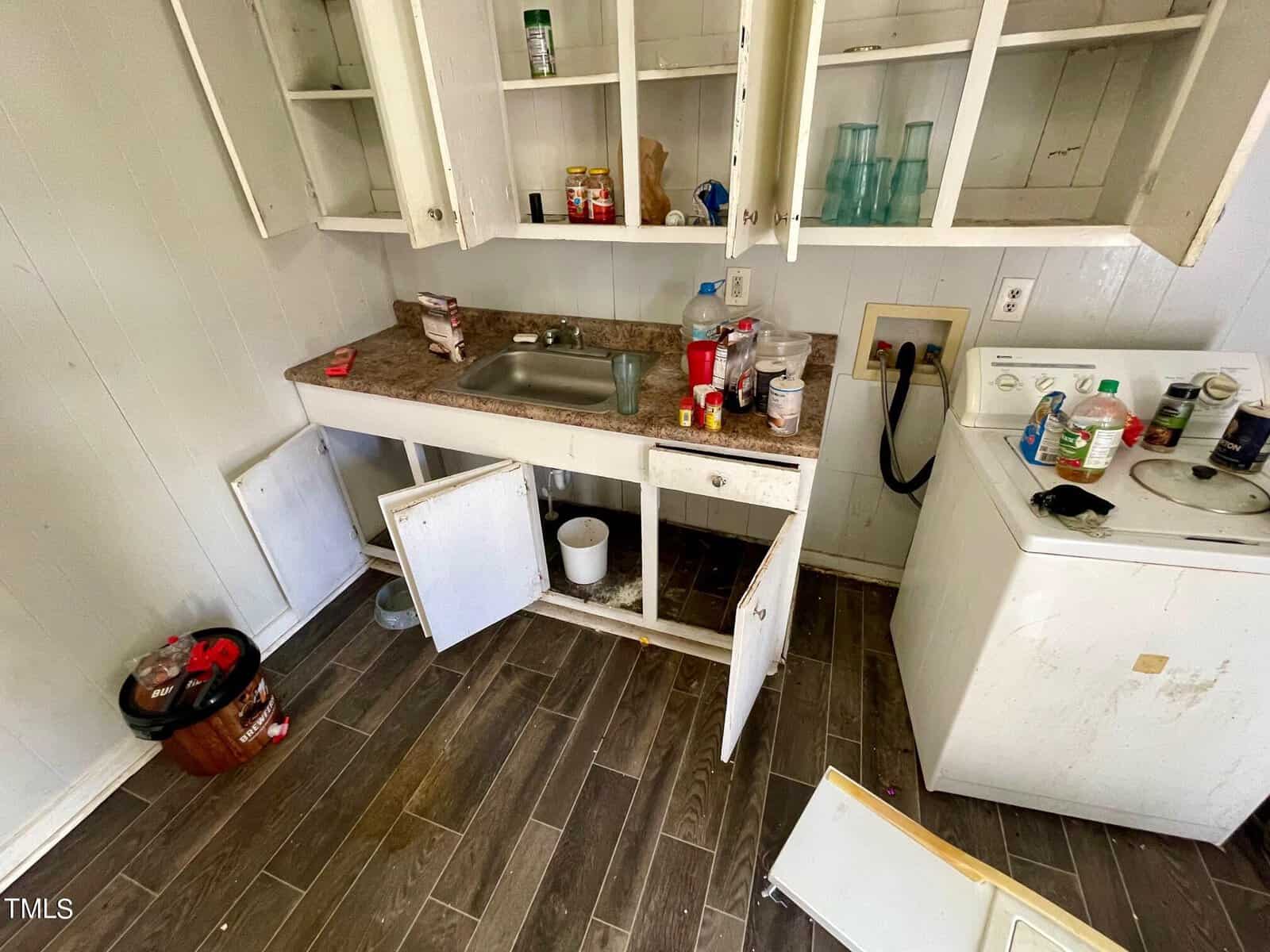 A cluttered and untidy kitchen area with open cabinets, a sink filled with dishes, and various items scattered on the countertops and floor.