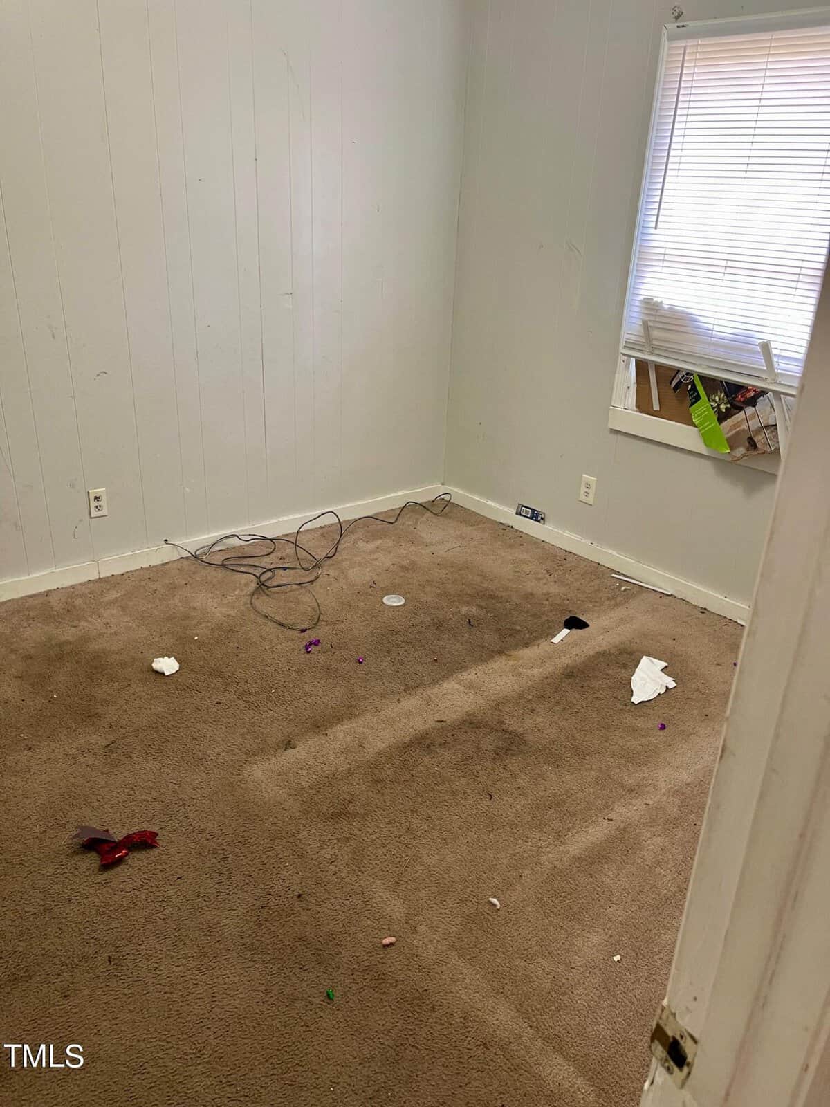 An empty room with stained carpeting and a few pieces of litter on the floor, with white walls in need of touch-ups and a window with a partially visible outside view.