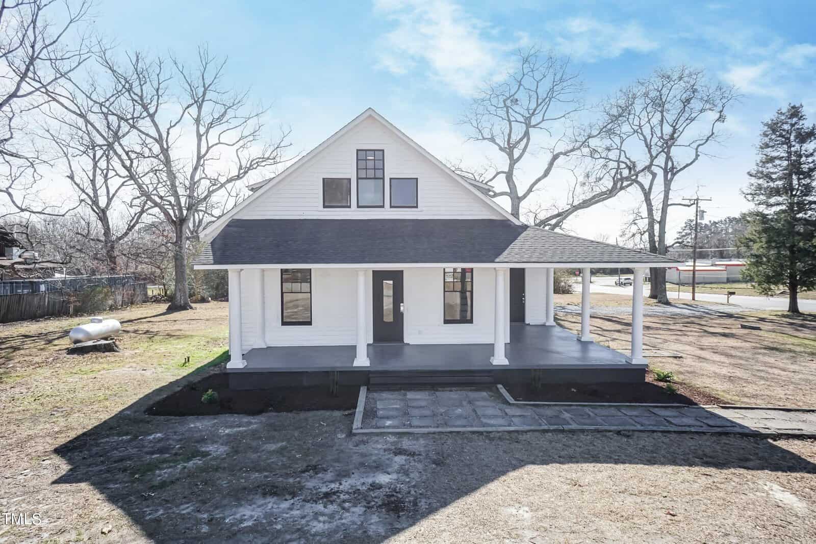 A white, two-story house with a gabled roof and a wraparound porch sits on a clear day. The property features a newly paved walkway, bare trees, and minimal landscaping. The sky is blue with some clouds.