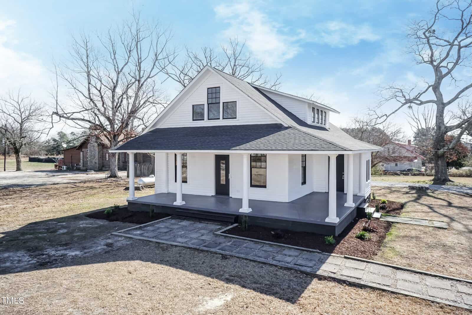 A white, two-story house with a large front porch, dark roof, and multiple windows stands on a grassy plot. Leafless trees surround the area under a clear, blue sky.