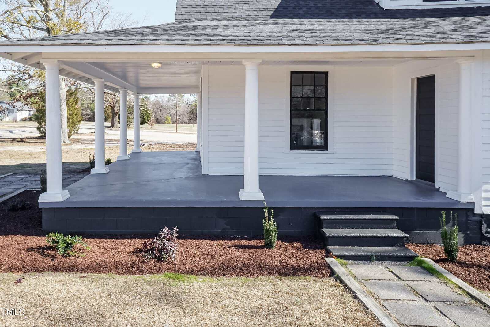A white house with a covered porch featuring several columns. The porch has steps leading up to a dark painted floor. Small plants are lined in a garden bed along the front. The surroundings include a lawn and trees in the background.