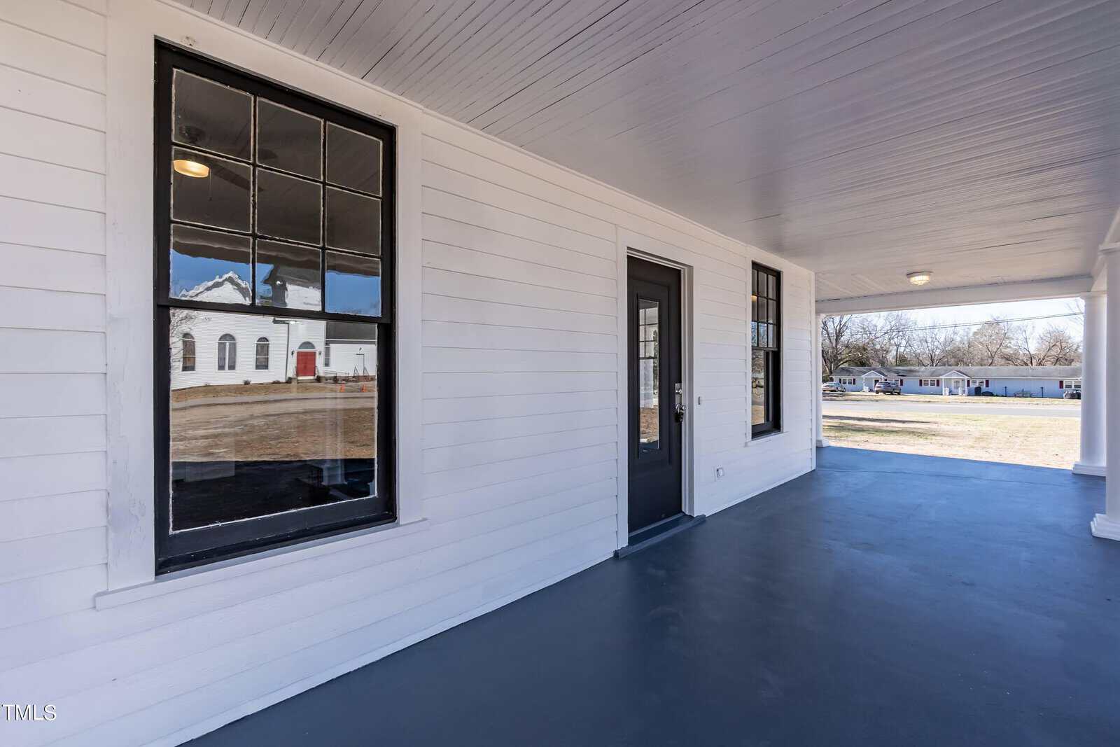 A spacious white porch with black-framed windows and a black door. The floor is painted in a dark gray color. In the reflection of the window, a white building with red accents is visible across the street.