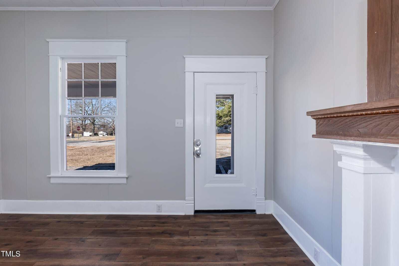 A small room with light gray walls and dark hardwood flooring. There is a window on the left and a door with a window in the center. Part of a white fireplace mantel is visible on the right. Outside, trees and grass can be seen.
