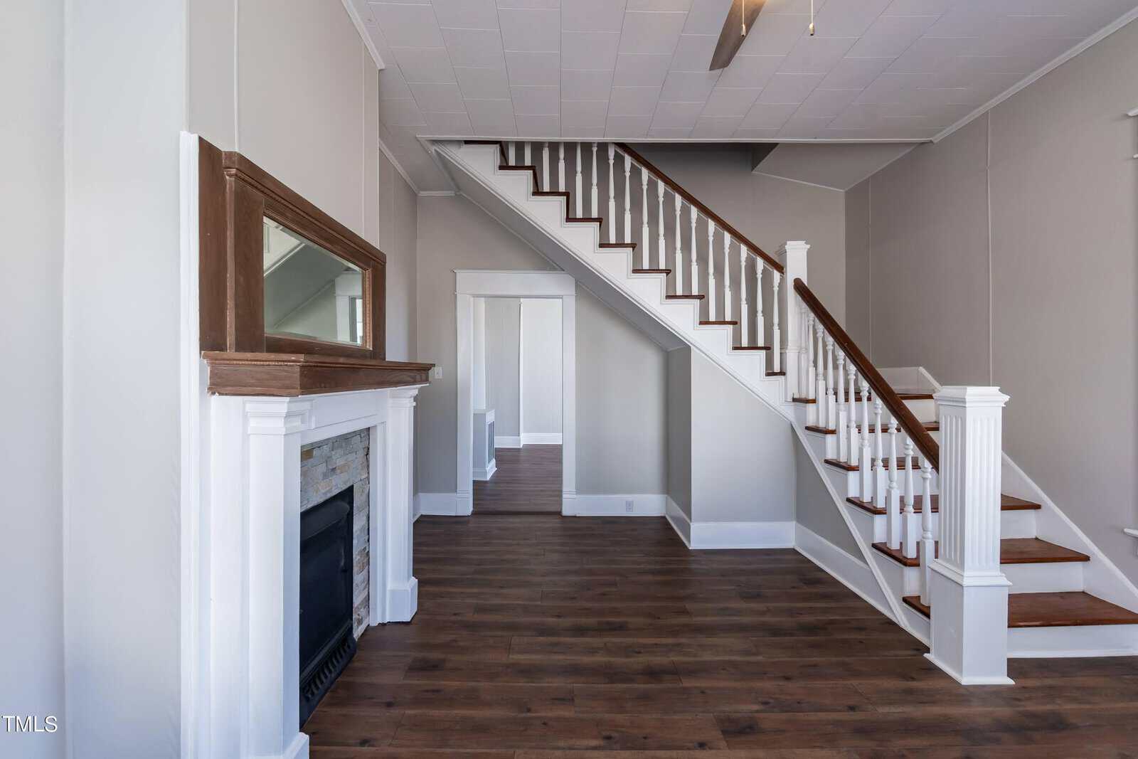 Living room with wooden floors, a fireplace with a wooden mantle, and a white staircase leading upstairs. The space is painted in light gray tones, and there's a large rectangular mirror above the fireplace.