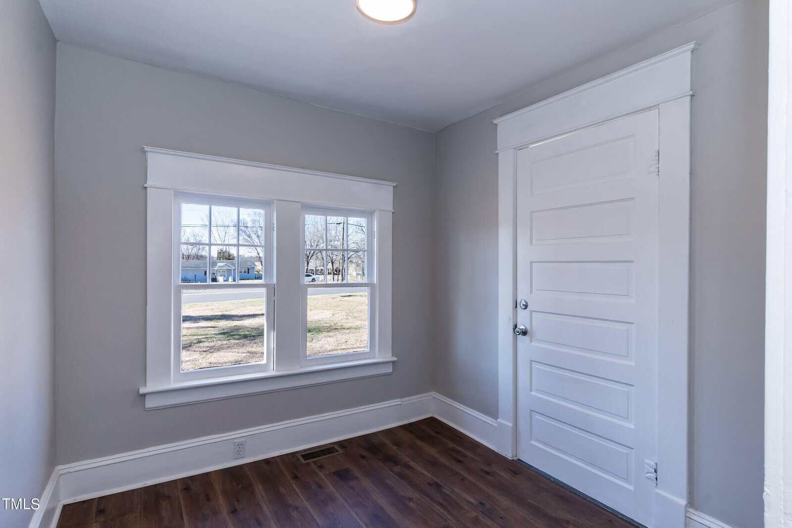 A small, well-lit room with light gray walls and dark wooden flooring. The room features two large windows that allow natural light to enter, and a white door with traditional molding.