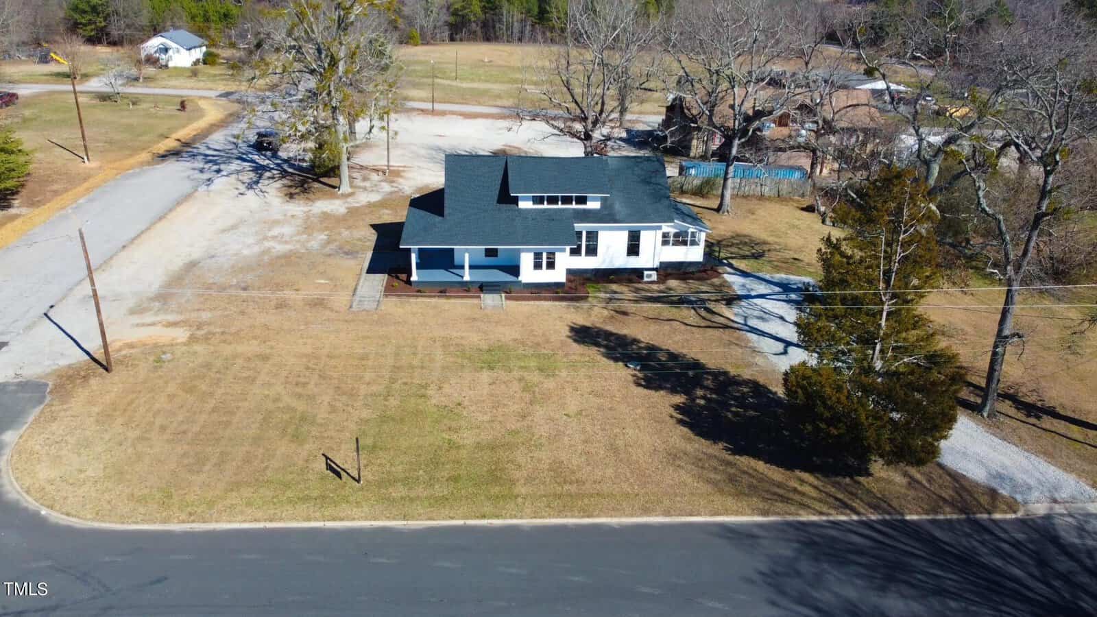 Aerial view of a blue single-story house with a dark roof, surrounded by a large grassy yard. A paved road runs in front, and there are a few leafless trees around the property. A small gravel driveway leads to the house.