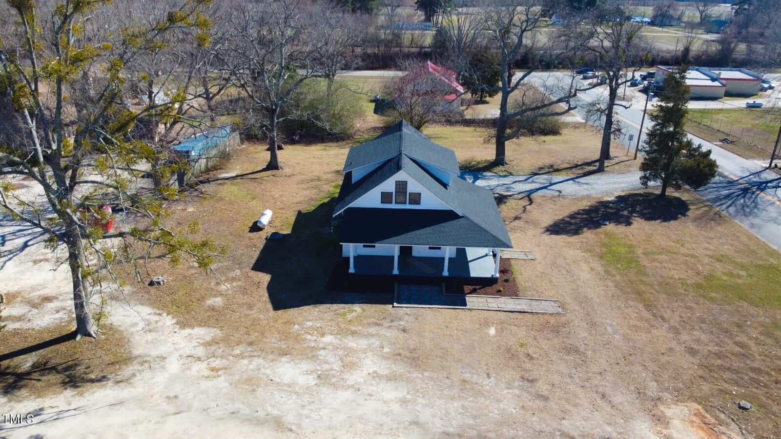 Aerial view of a two-story house with a dark roof, surrounded by bare trees and vast open land. The house has a wraparound porch and a driveway leading to a nearby road. The landscape is lightly dusted with snow.