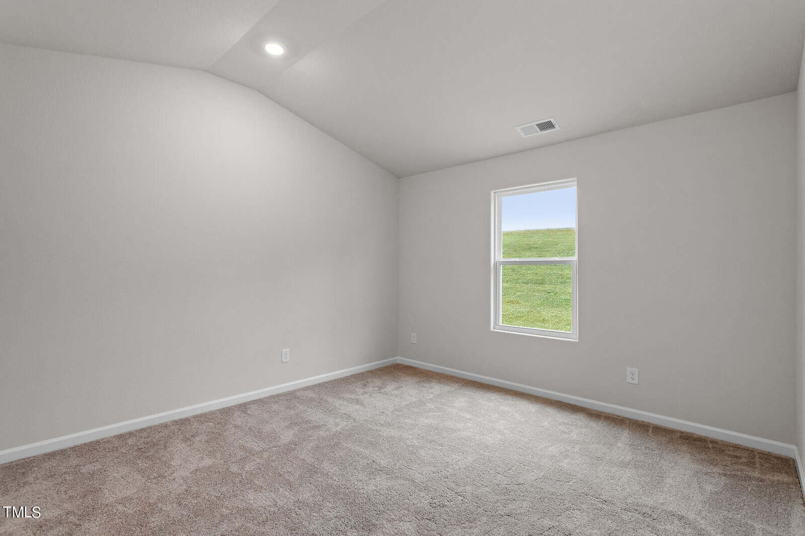 An empty room with beige carpet and light gray walls. The ceiling is vaulted with a recessed light. A single window shows a view of green grass outside. No furniture or decorations are present.