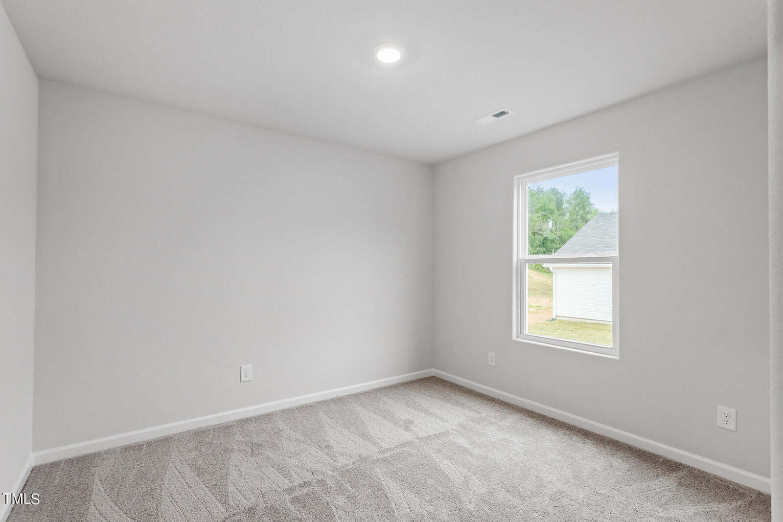 A small, empty room with light gray walls and beige carpet. A single window lets in natural light, showing a view of a white building and greenery outside. The ceiling has a recessed light, and electrical outlets are visible on the walls.
