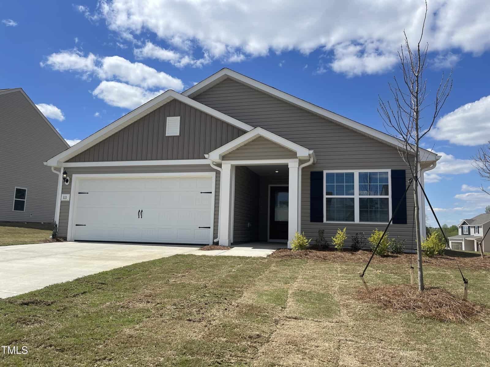 Nestled on 111 Cultivar Street, this modern single-story house in Oxford boasts gray siding and a pristine white garage door. A newly planted tree graces the front yard alongside lush patches of grass, all beneath a clear sky with just a few clouds.