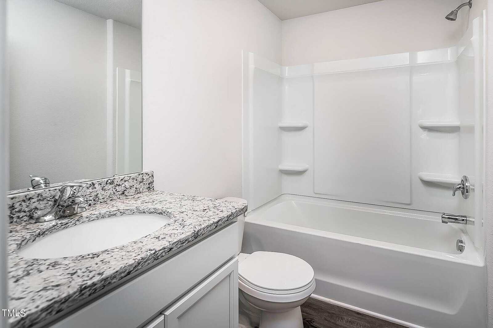 A modern bathroom with a granite countertop vanity, a white sink, chrome faucet, and a toilet. Adjacent is a white bathtub with a shower, surrounded by a smooth white wall. The floor has light-colored wood flooring.