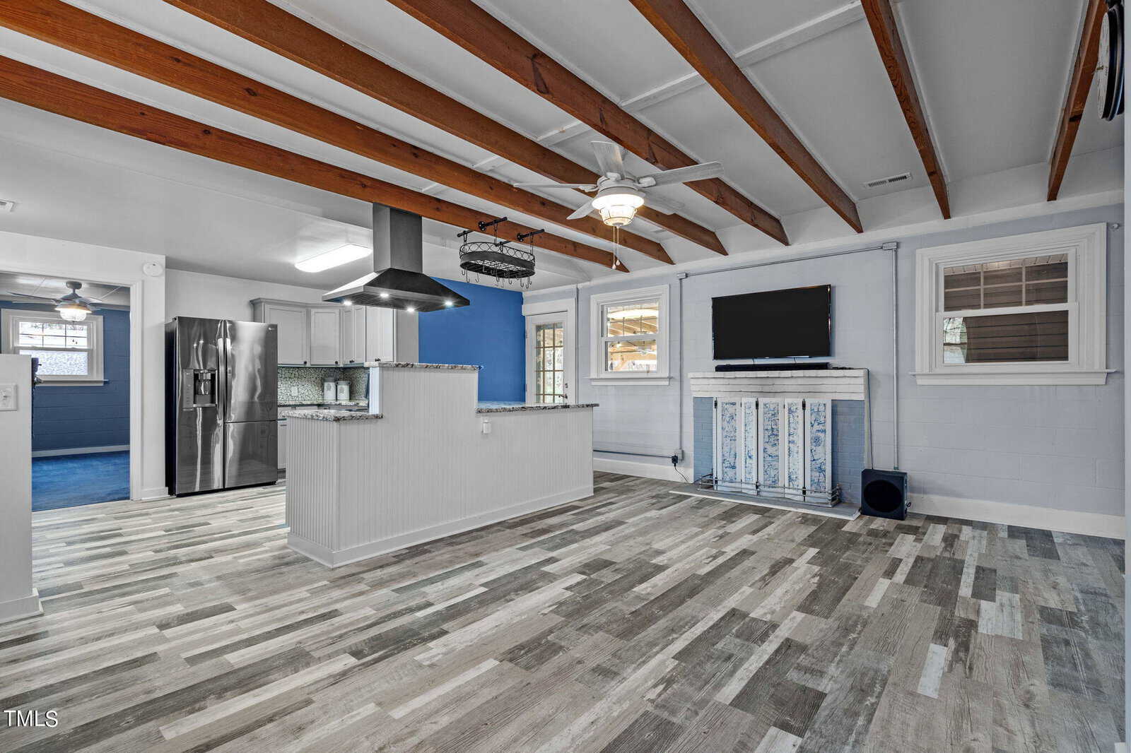 A modern kitchen and living space with exposed wooden beams, light blue walls, and light wood flooring. The kitchen features a stainless steel refrigerator, sleek countertops, and a range hood. A TV is mounted above a decorative mantle.
