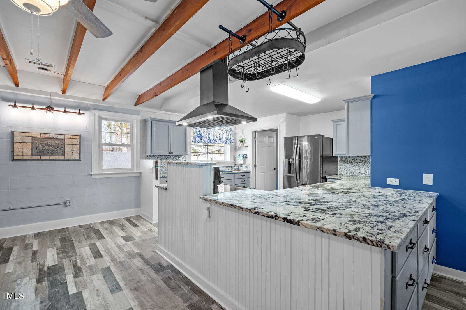 A modern kitchen features exposed wooden beams, a granite countertop island, light blue cabinets, stainless steel appliances, and a ceiling fan. The walls are light gray, and there is a large window above the sink and a narrow brick-colored backsplash.