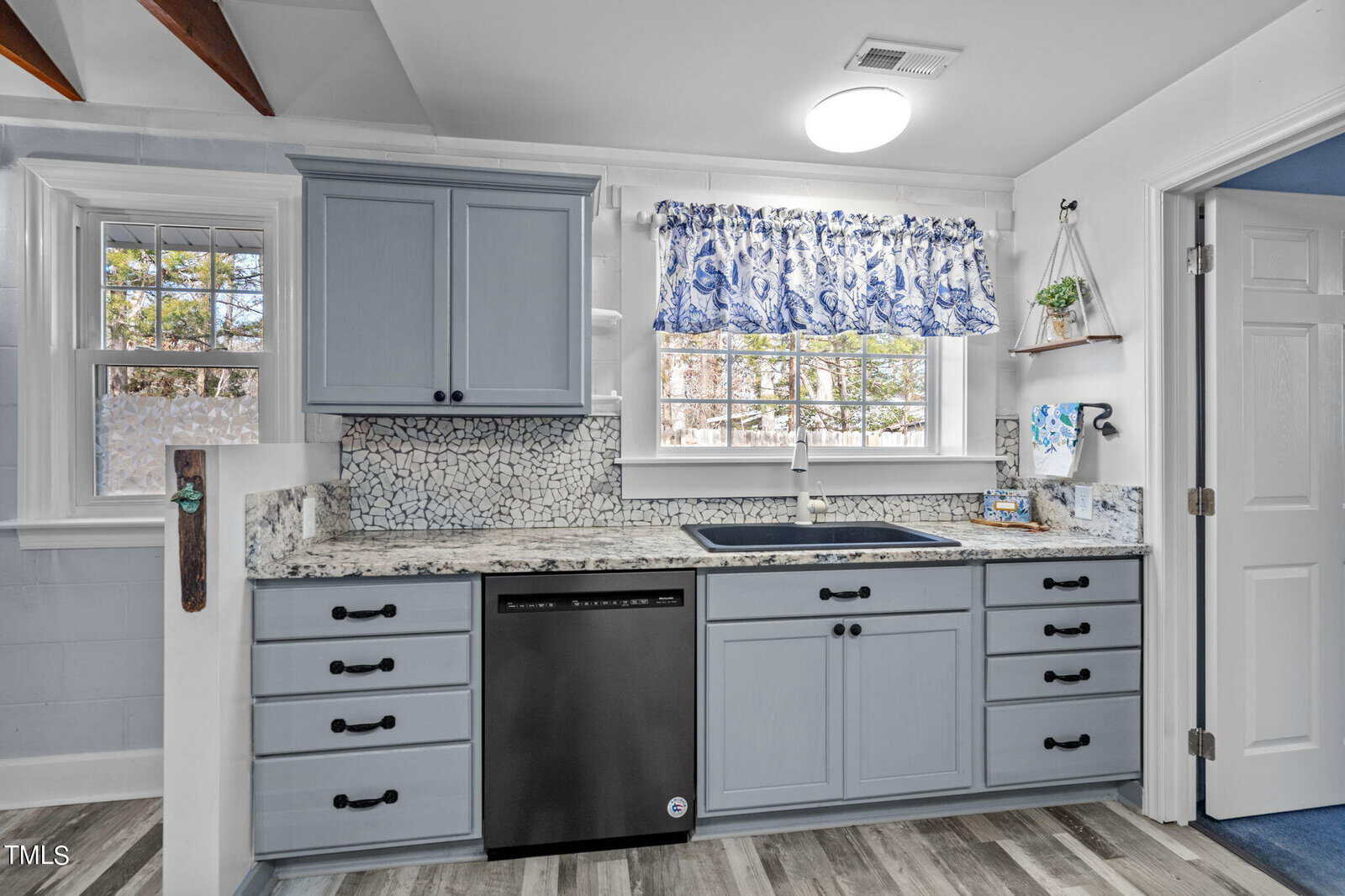 A modern kitchen with light blue cabinets, a speckled granite countertop, and a black dishwasher. A window with a blue and white patterned curtain is above the sink. The floor is wood, and the walls are white.