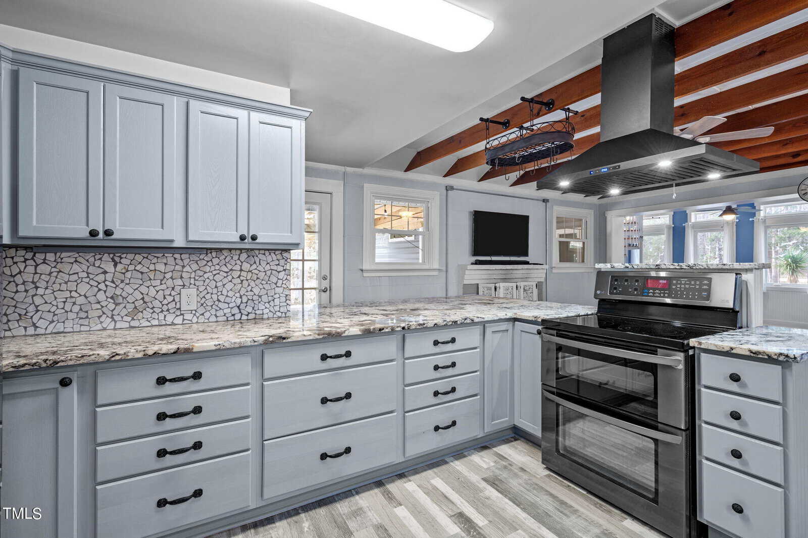 A modern kitchen with light blue cabinets, granite countertops, and an electric stove. The room features overhead wooden beams, a mosaic tile backsplash, and a large window, creating a bright and inviting space.