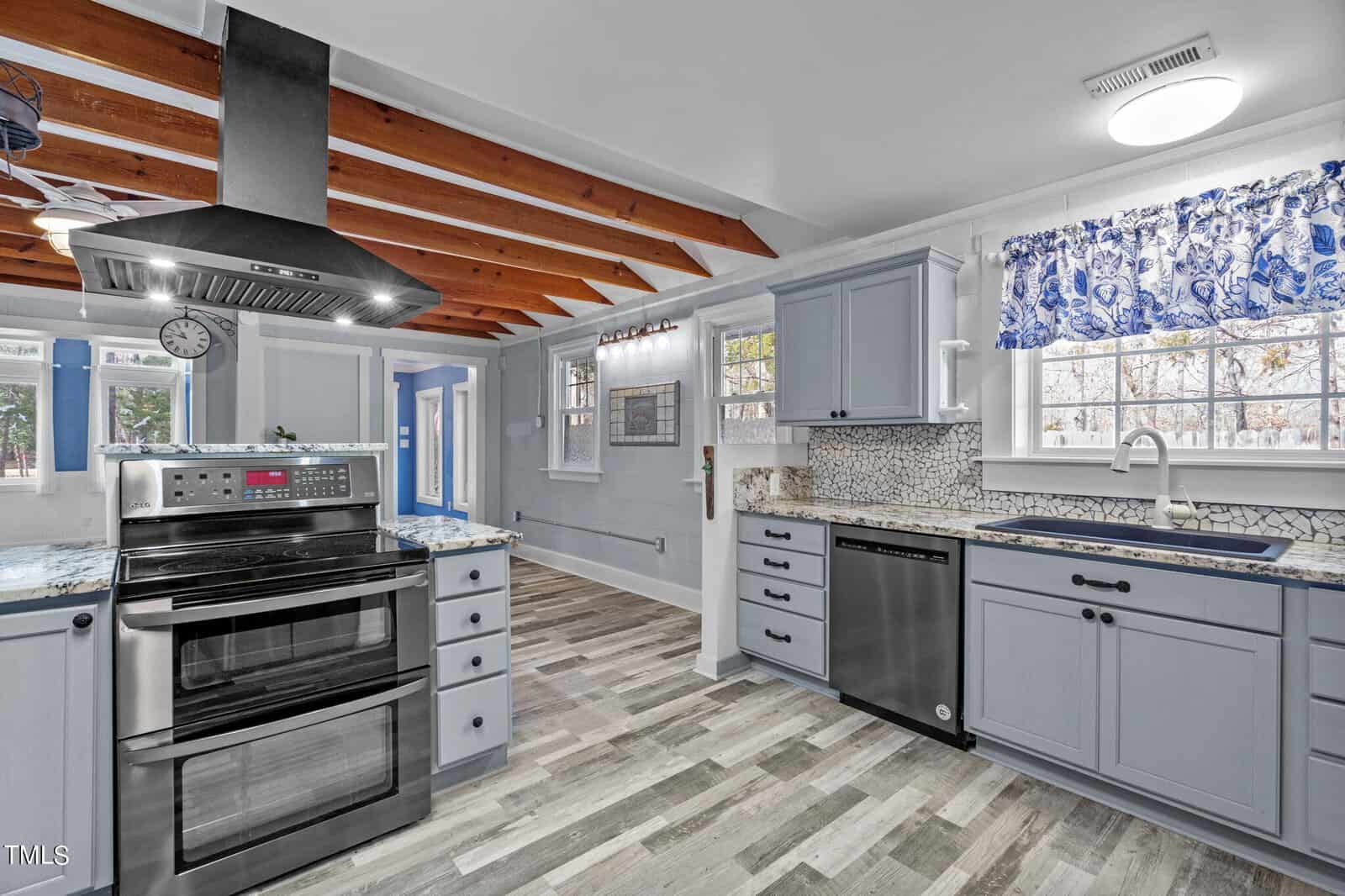 A modern kitchen with light blue cabinets, a double oven, stainless steel appliances, and a mosaic tile backsplash. The space features a wooden ceiling beam, patterned curtain above the sink, and a clock on the wall.