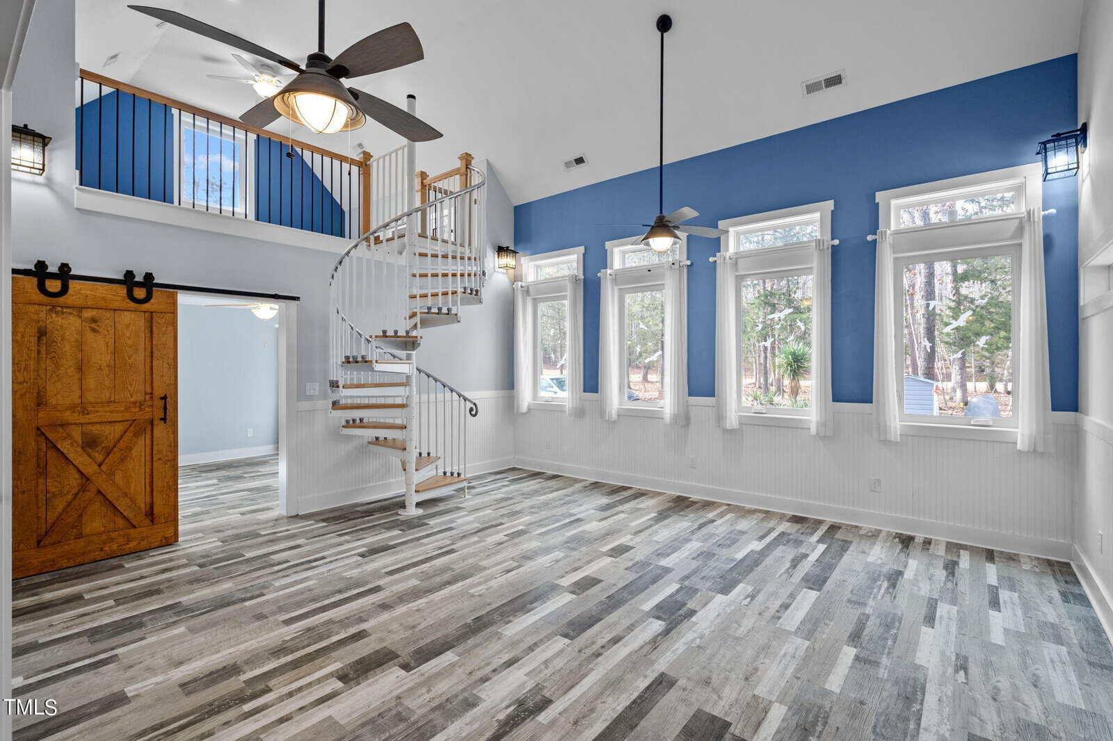 A spacious living room with a spiral staircase, high ceiling, and five large windows. The room features a blue accent wall, wood-look flooring, ceiling fans, and a sliding wooden barn door on the left, leading to another room.