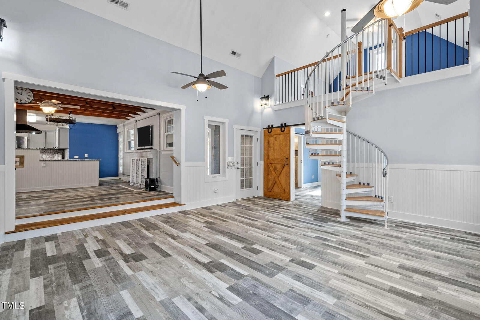 A spacious interior featuring a two-tone gray wood floor, light blue walls, and a vaulted ceiling. A spiral staircase leads to an upper level. The room is open-plan with a visible kitchen area, wooden beams, and modern lighting fixtures.