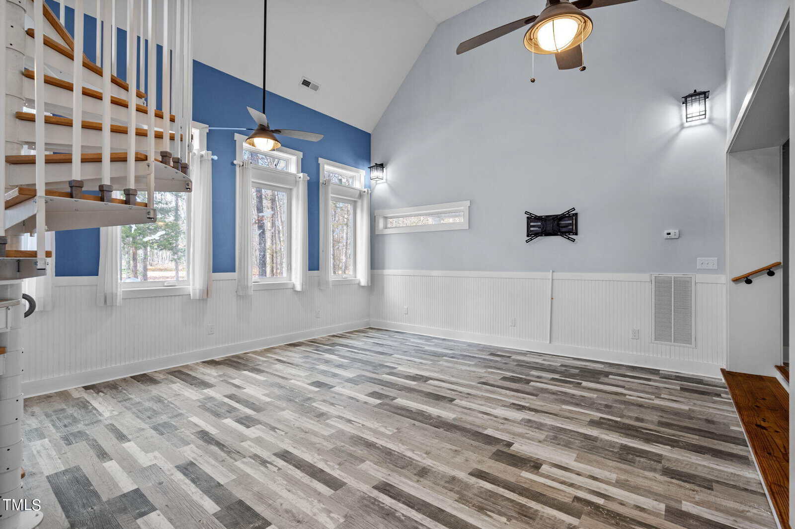 A spacious room with high ceilings, light blue and white walls, and wood-patterned flooring. Large windows allow natural light. A curved wooden staircase leads upwards, and there are wall-mounted lights and a space for a wall TV.